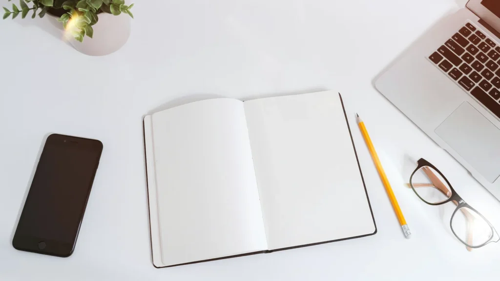 An open notebook on a white desk next to an iPhone and a MacBook