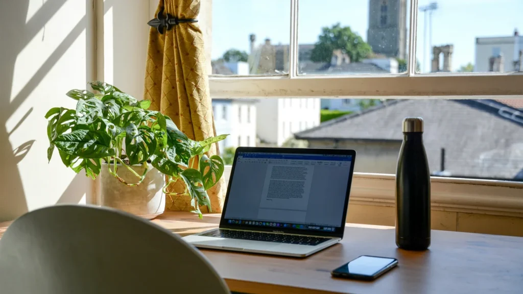 A laptop on a desk by a sunny window overlooking rooftops
